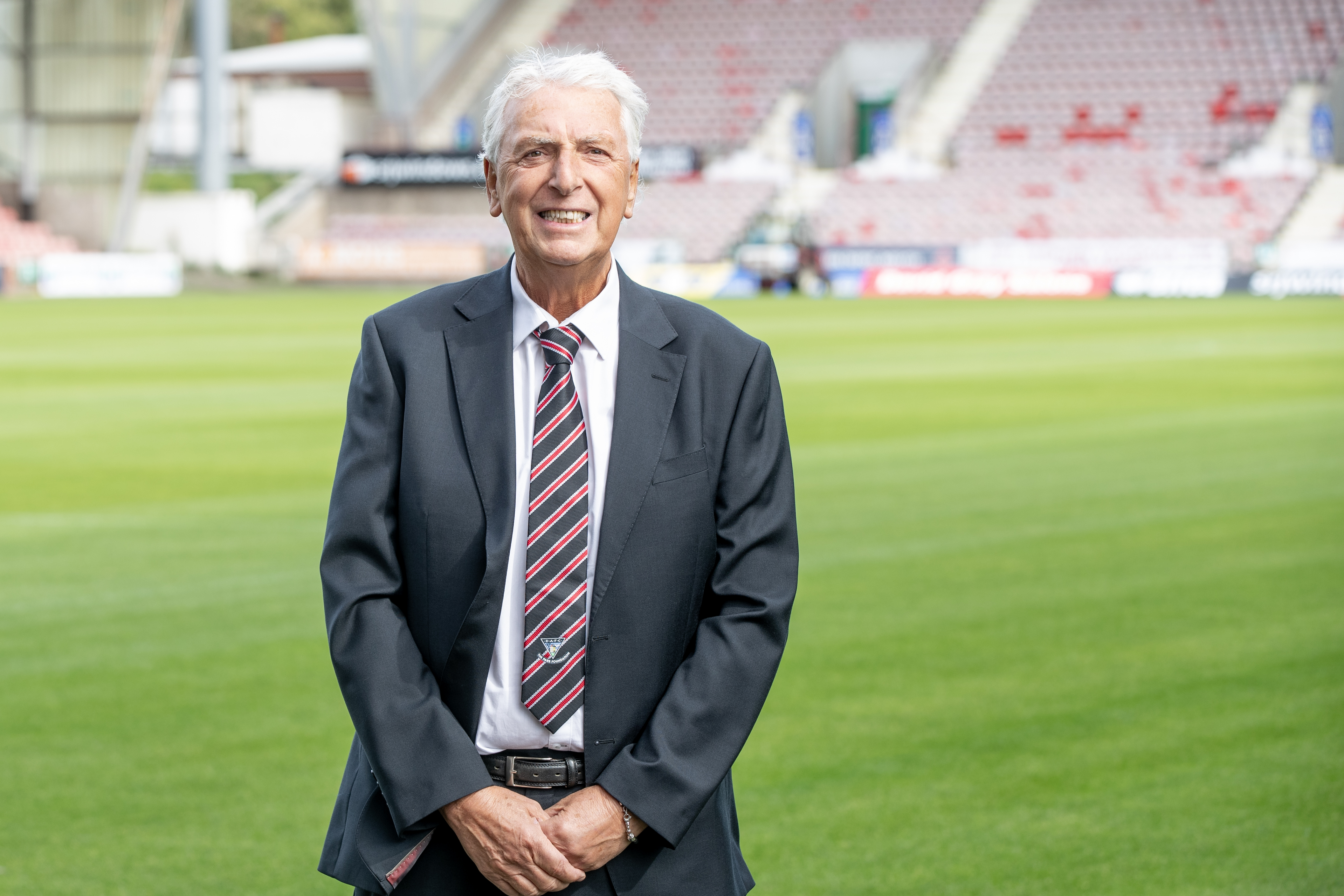 Dave, who is wearing a suit, is smiling at the camera while standing on a football pitch