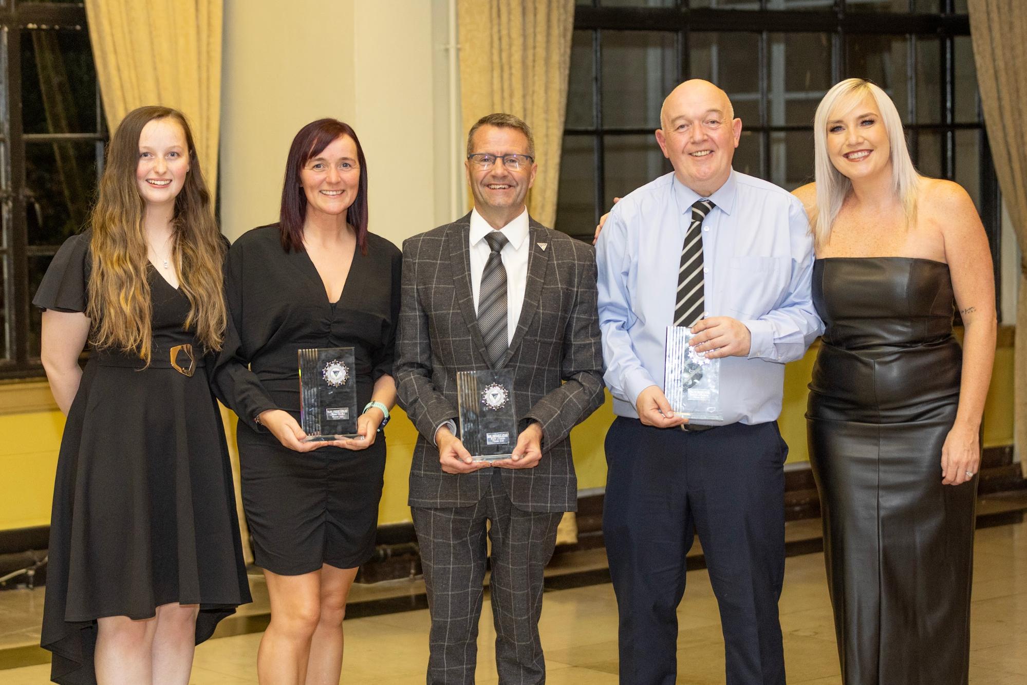 Five people stood, with three of them holding awards while smiling at the camera