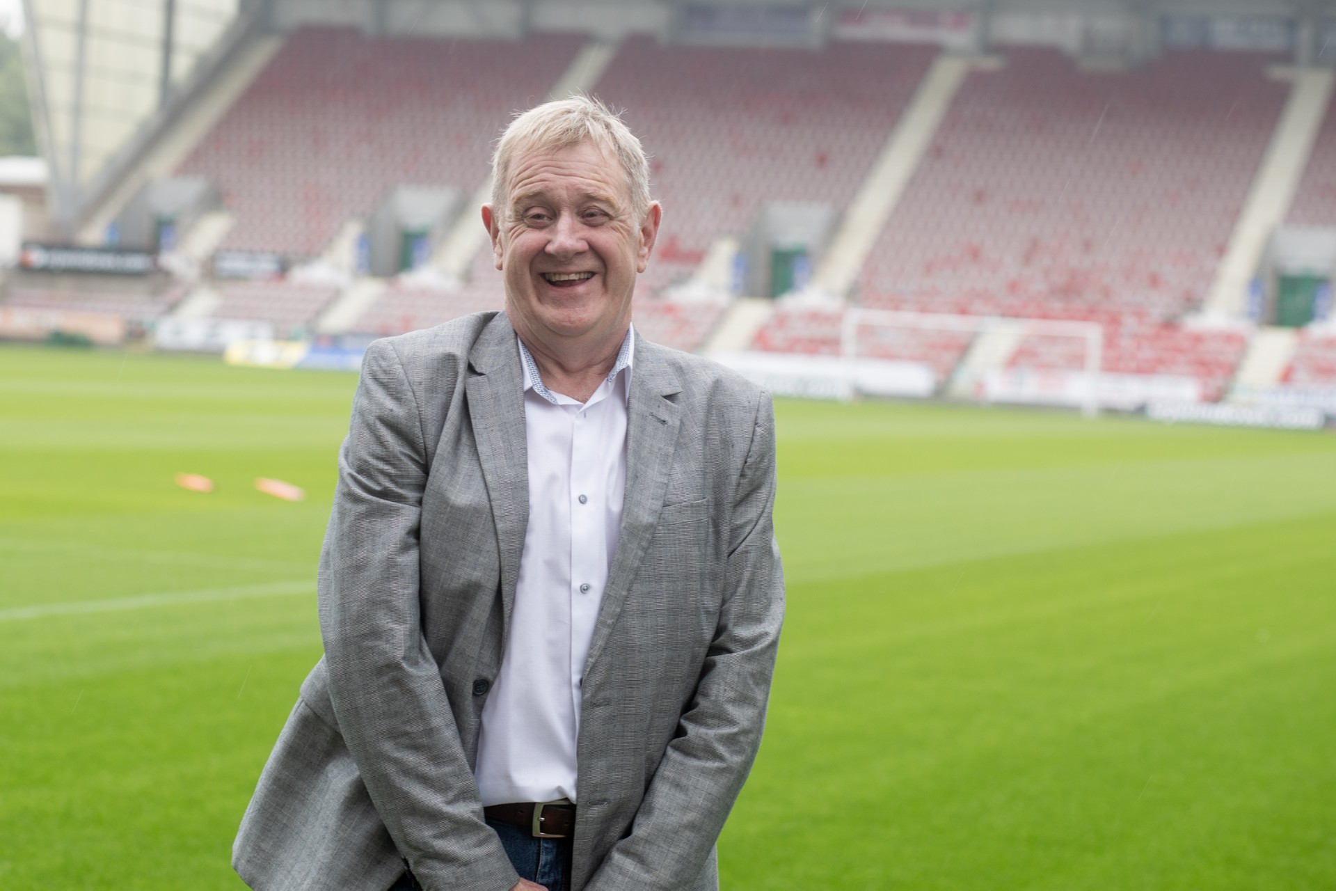 Tony Combe wearing a white shirt and a grey blazer inside a stadium smiling