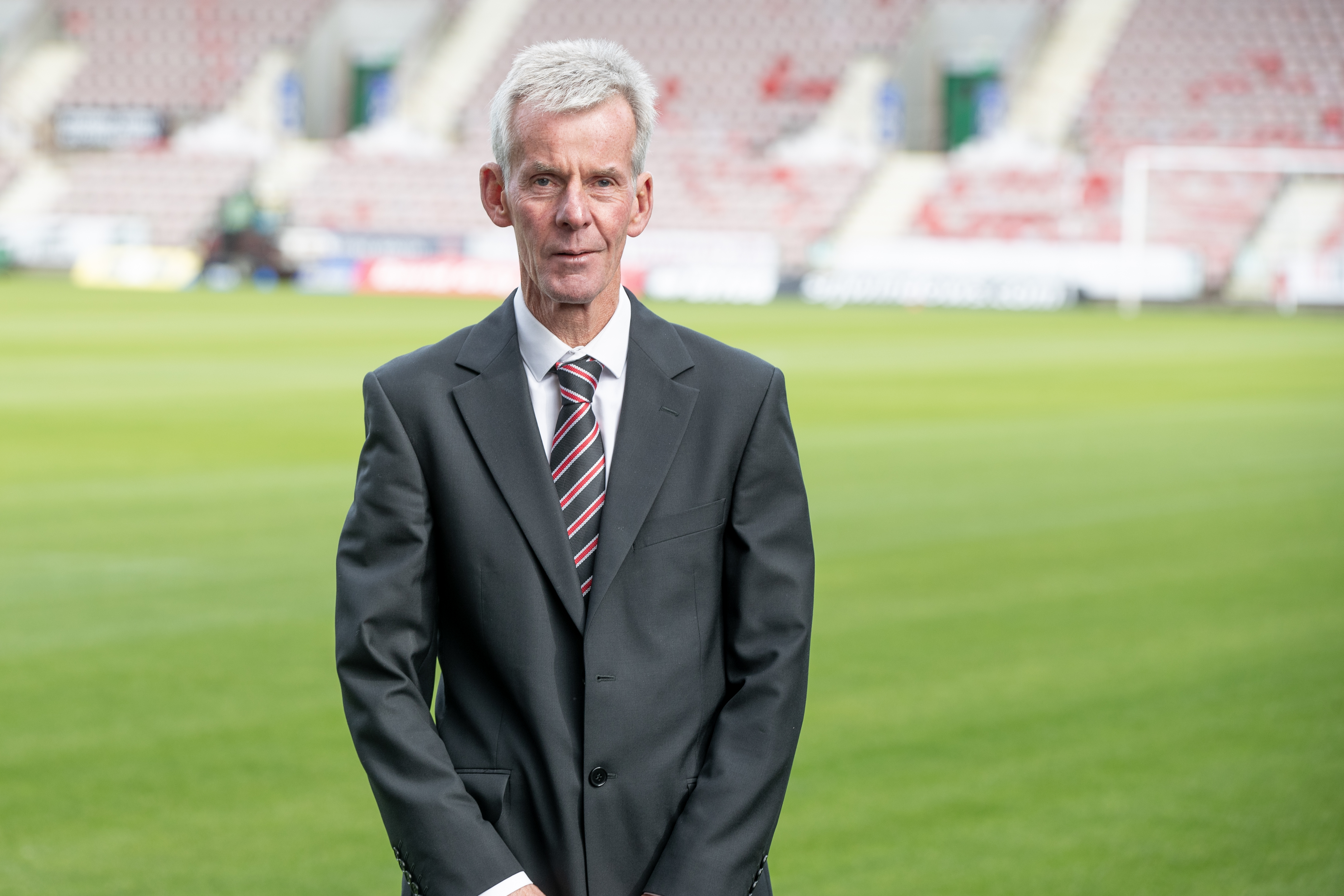 Alexander, wearing a suit, is smiling at the camera while standing on a football pitch