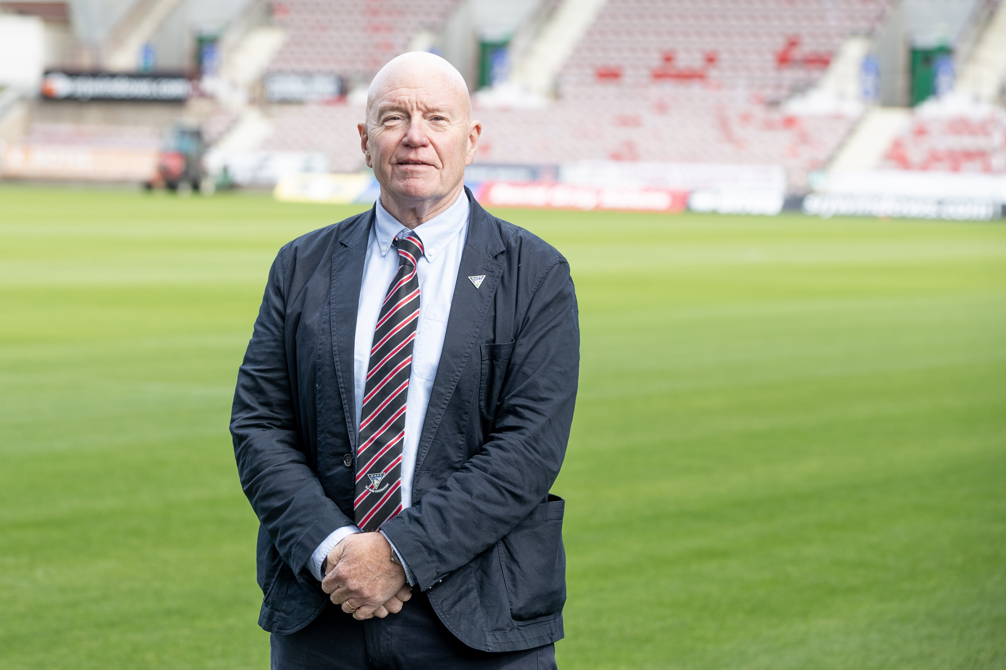 Derek, wearing a suit, is smiling at the camera while standing on a football pitch