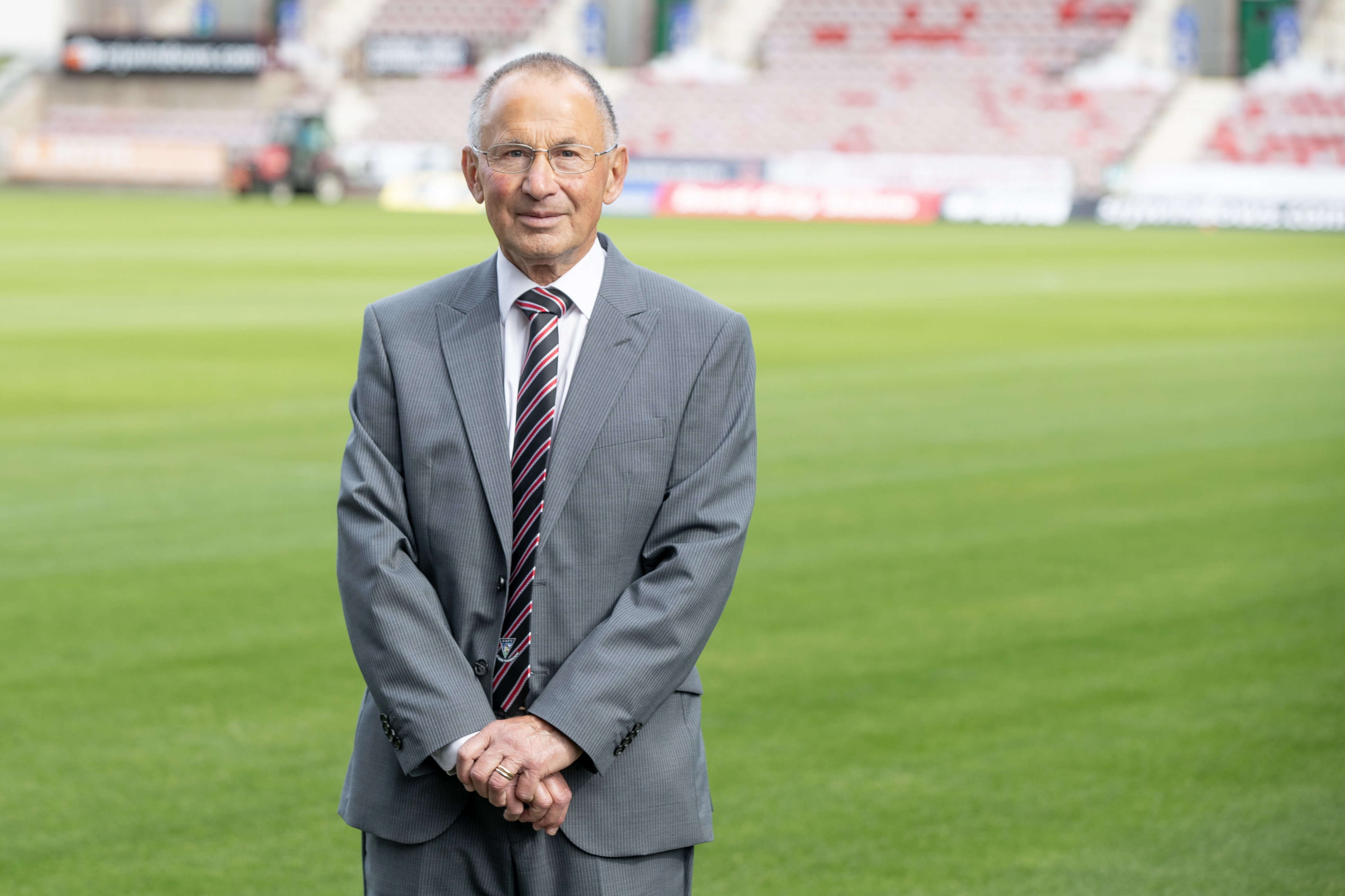 Gennaro, wearing a suit, is smiling at the camera while standing on a football pitch