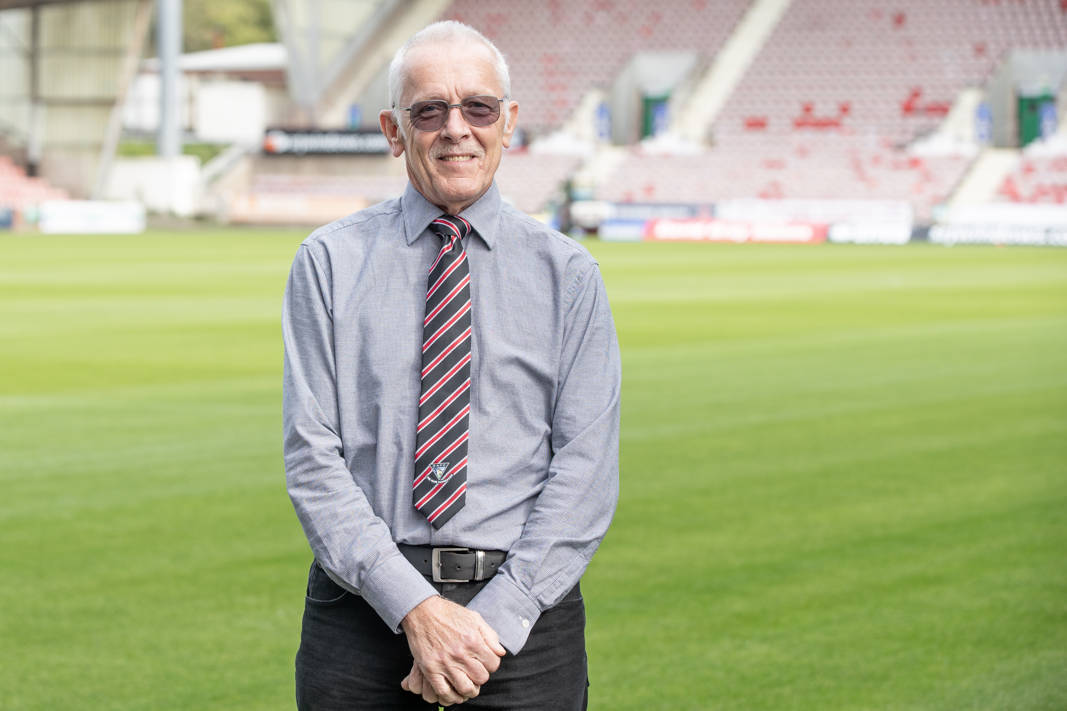 Alan, wearing a suit, is smiling at the camera while standing on a football pitch